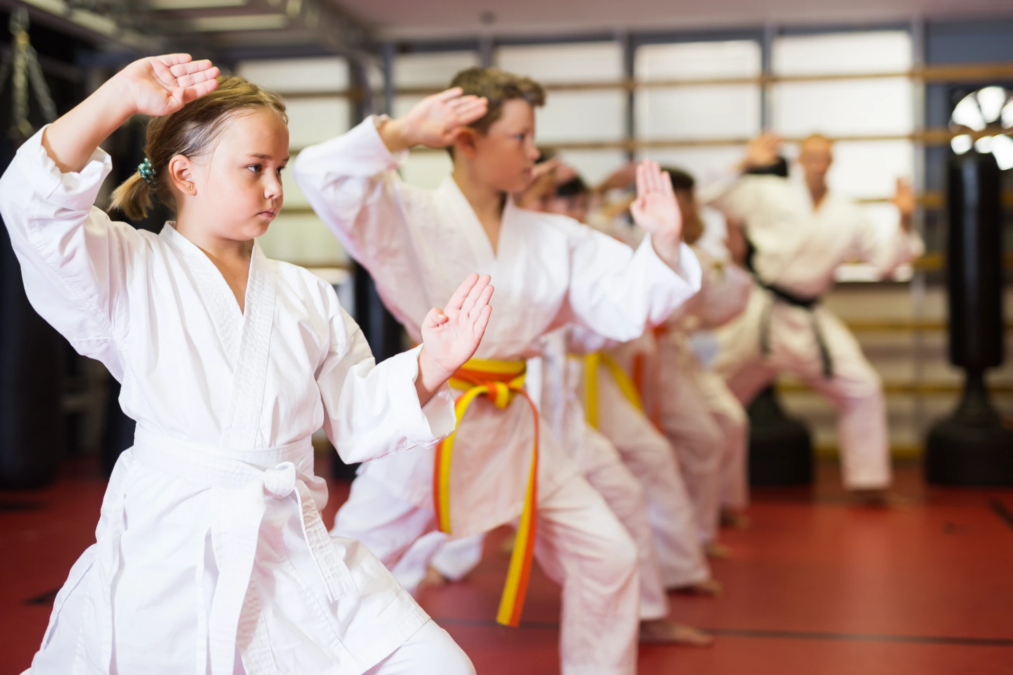 A line of students in white karate gis, including a focused young girl in the foreground, practice a high block stance, led by a black belt instructor.