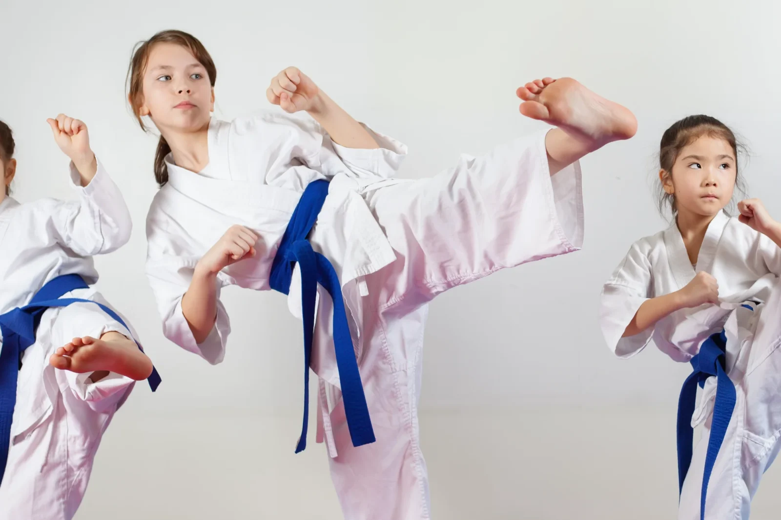 Three youth girls wearing white karate uniforms and blue belts practice performing martial arts kicks with serious, focused expressions.