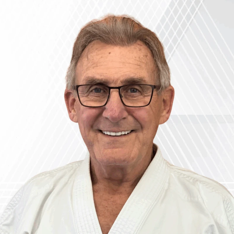 Shihan Randy Holeman, an older man wearing glasses and a white karate gi, smiles broadly in a professional headshot.