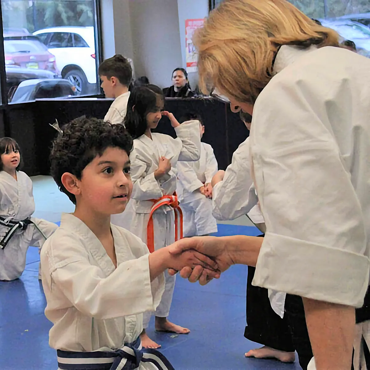 A young boy in a blue-belted white gi shakes hands with his instructor in a martial arts studio. He looks up with a proud expression while other students stand behind him.