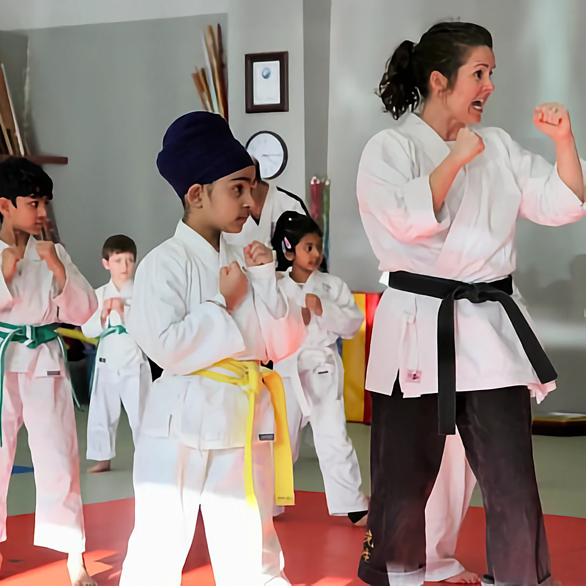 A focused black belt instructor demonstrates a fighting stance to a group of young students with yellow and green belts during a karate class.