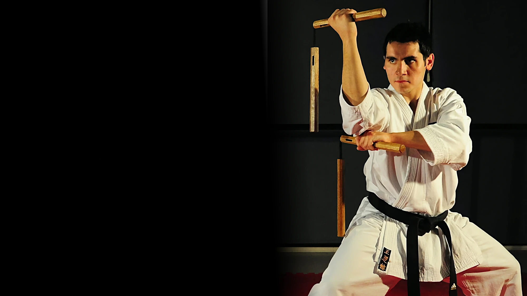 A focused male student in a white gi and black belt trains with two pairs of nunchaku, holding them in a ready stance against a dark background.
