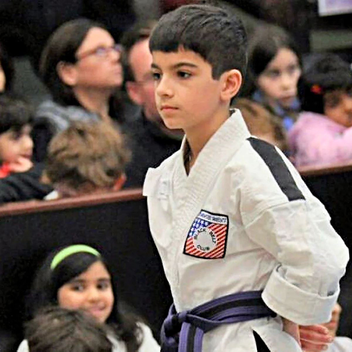 A youth wearing a white karate uniform and purple belt stands with a focused expression, hands behind his back, during a tournament.