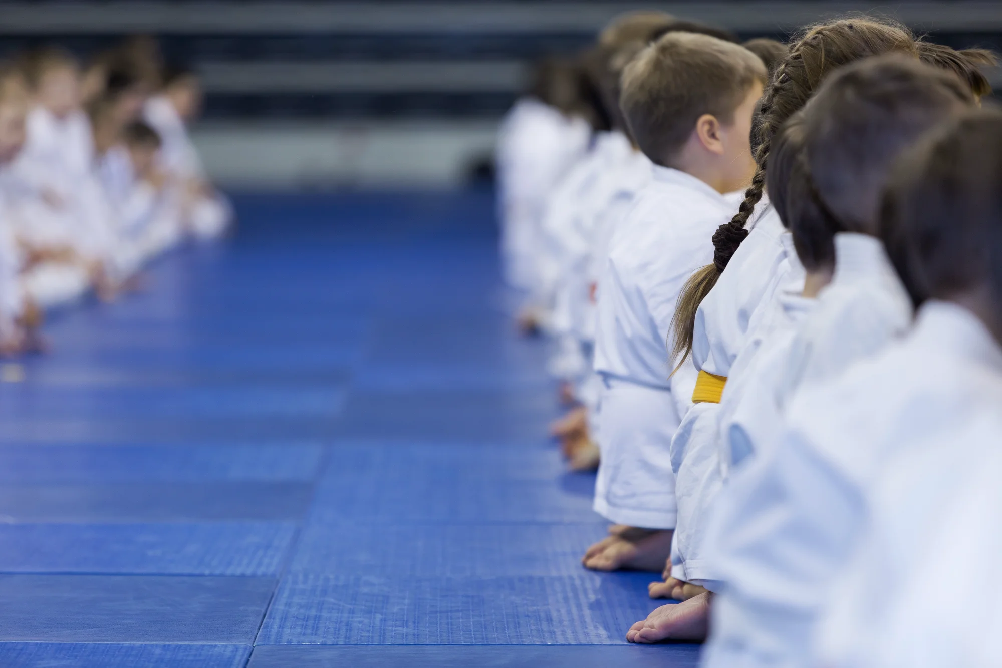 Young students in white gis and yellow belts sit attentively in two parallel rows on a blue martial arts mat, preparing for their class.