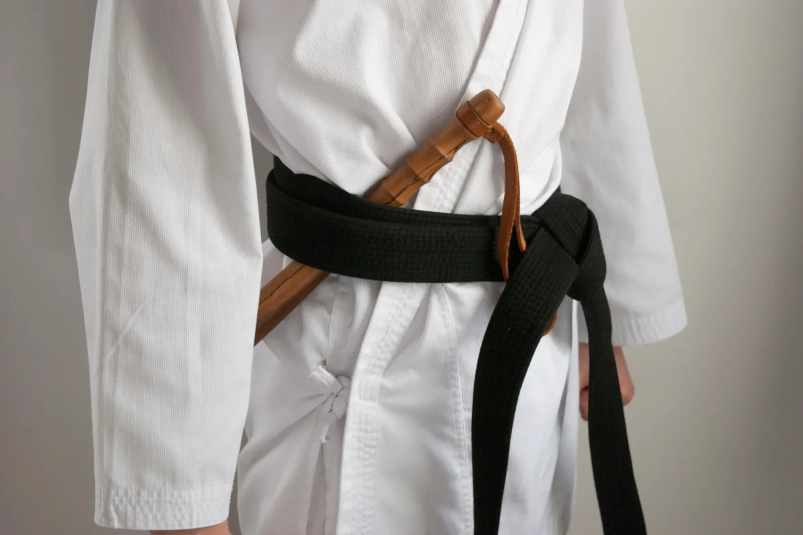 The torso of a martial artist wearing a white gi and a black belt. A wooden training weapon, visible beneath the belt, rests against the uniform.