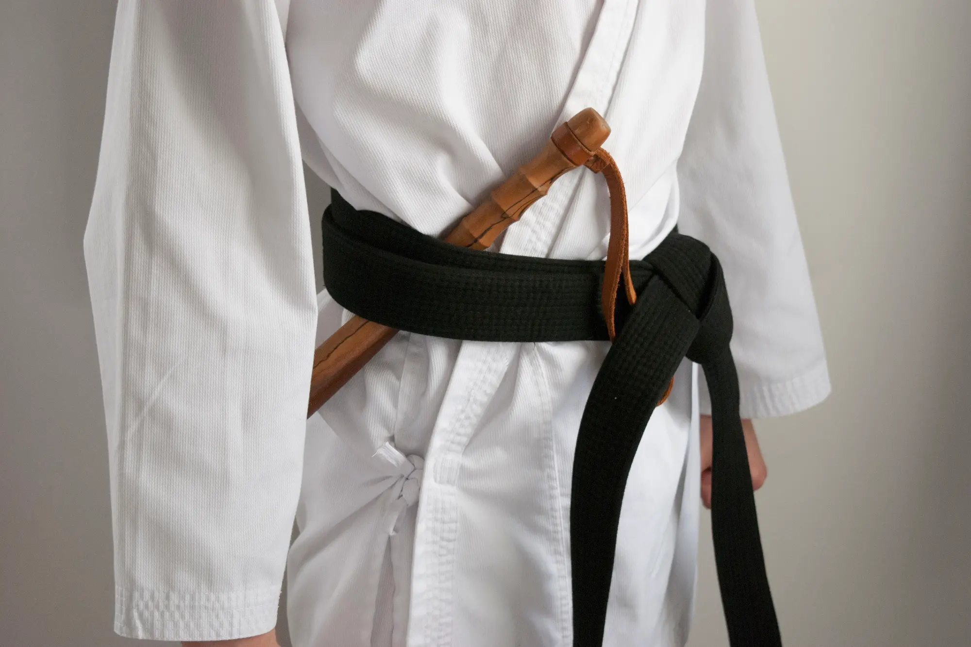 The torso of a martial artist wearing a white gi and a black belt. A wooden training weapon, visible beneath the belt, rests against the uniform.