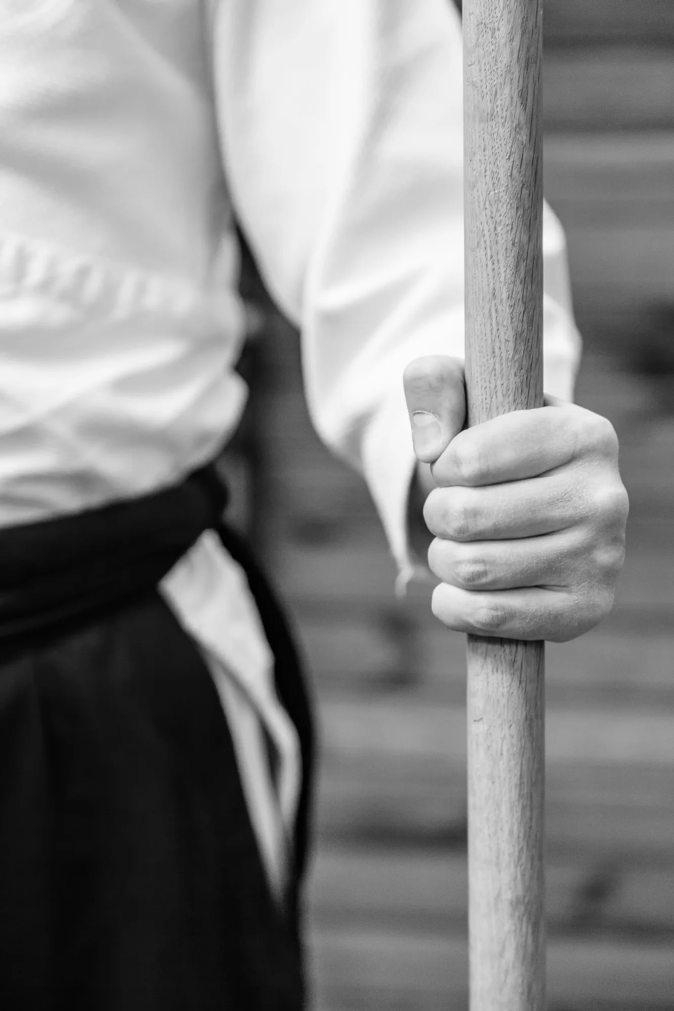 A person in a traditional martial arts uniform holds a wooden weapon staff in a tight hand grip.