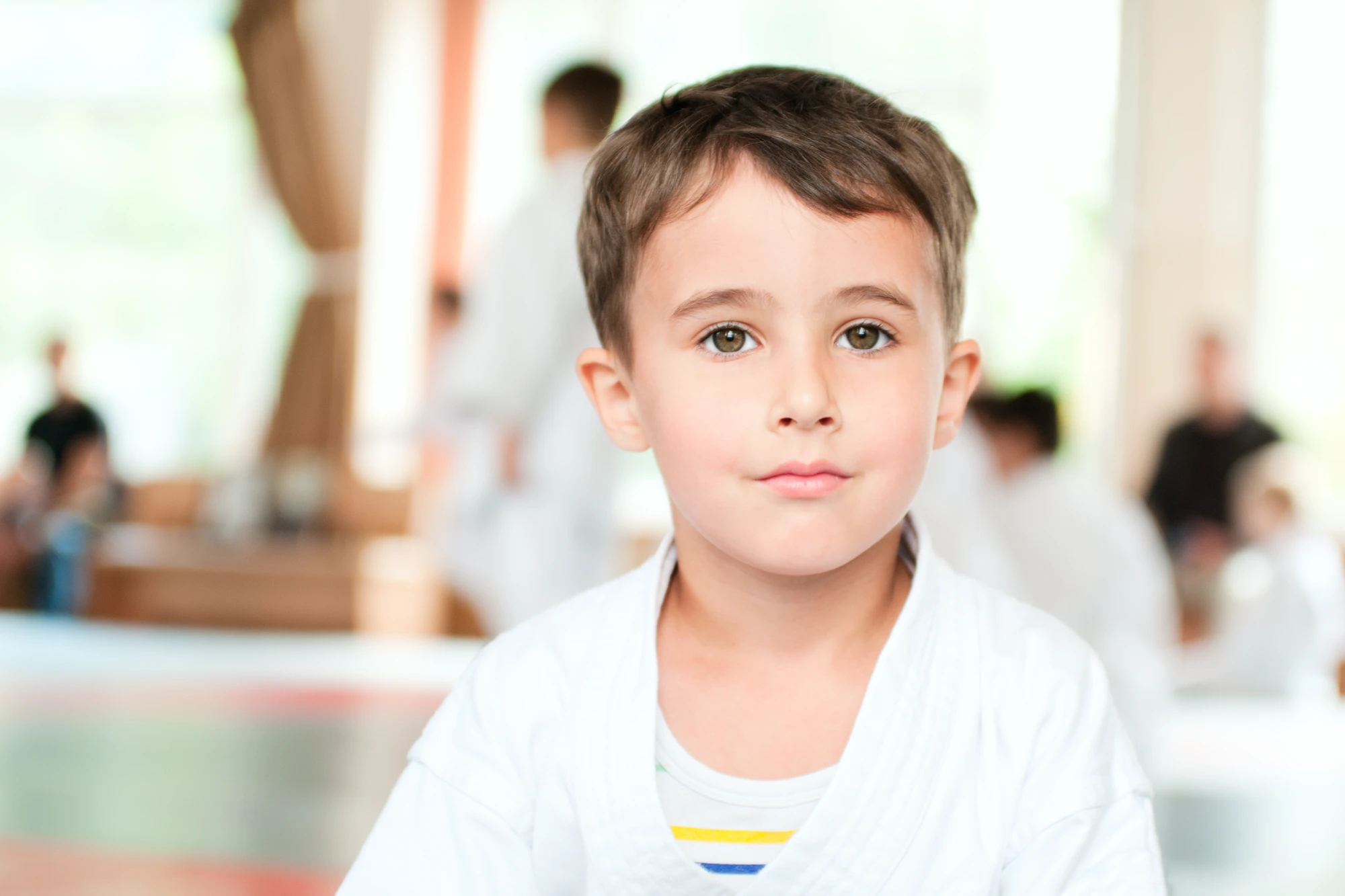 A young boy in a white karate gi looks directly at the camera with a focused expression. Other students are blurred in the background of the dojo.