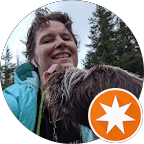 A person with short hair smiles widely while standing outside in a forest setting. A shaggy dog is joyfully licking the person's cheek.