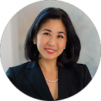 A professional portrait of a woman, Suzanne S., wearing a black business suit and a pearl necklace, smiling confidently at the camera.