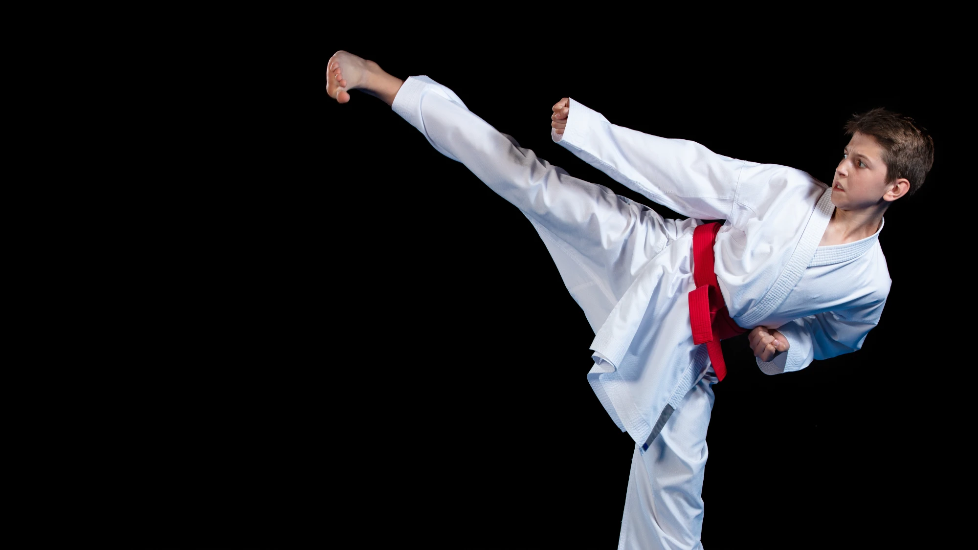 A young boy with a focused expression, wearing a white uniform and red belt, performs a powerful high kick.