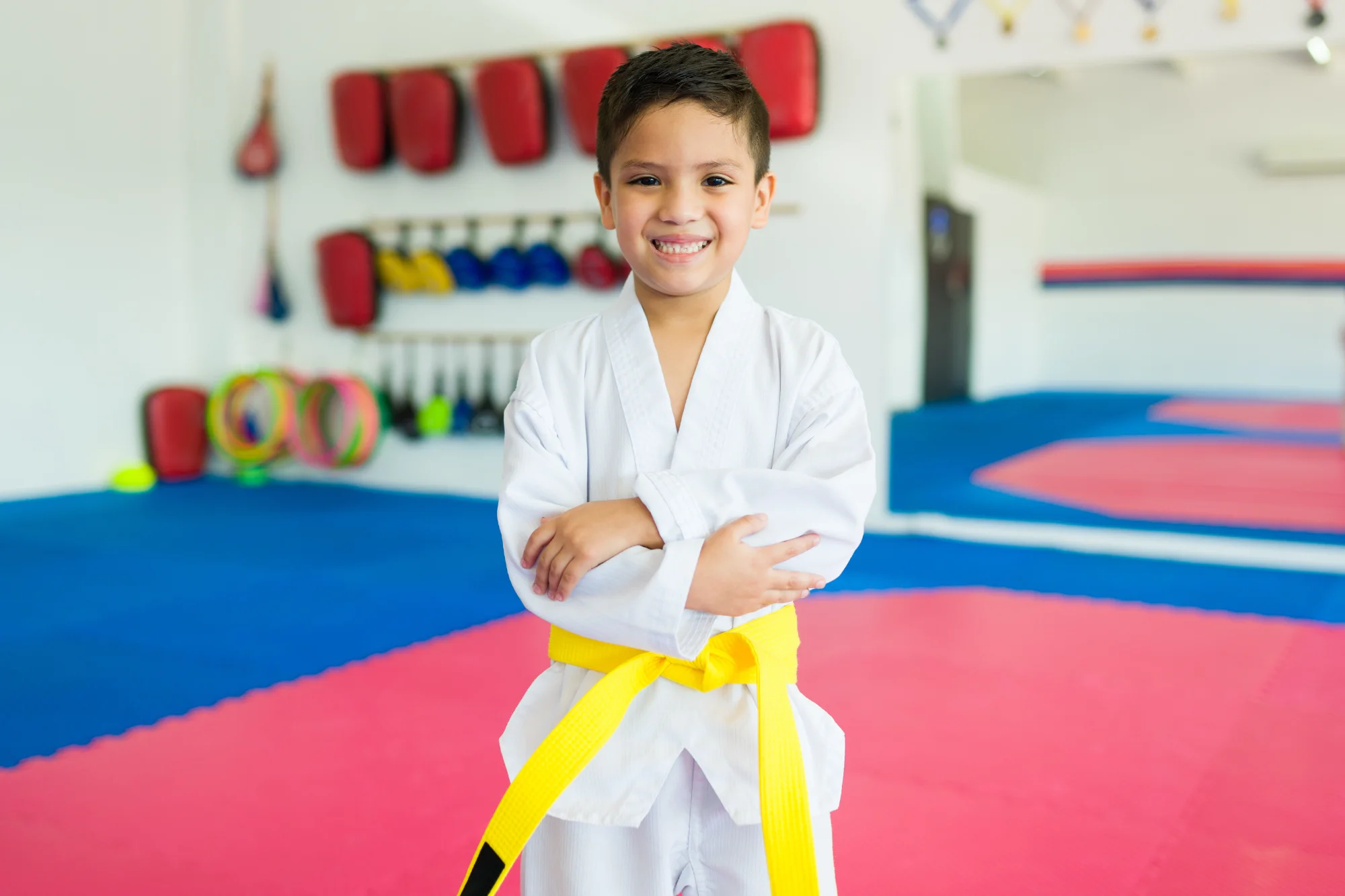 A young boy in a white karate gi and yellow belt stands smiling with arms crossed in a dojo setting with red and blue mats.