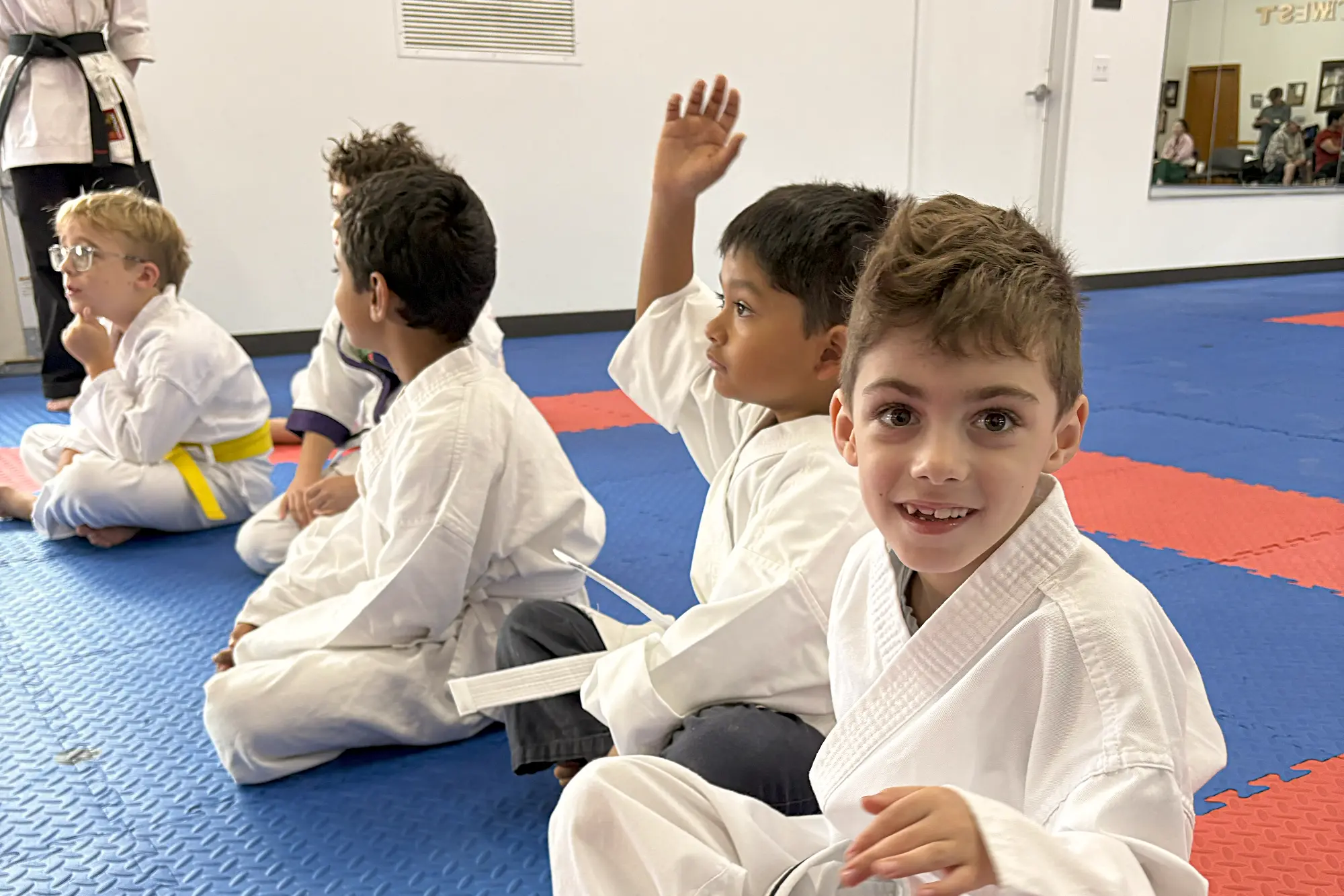 Several youth karate students in white gis sit on a blue mat. One boy with a yellow belt watches intently, another raises his hand, and a boy in the foreground smiles.