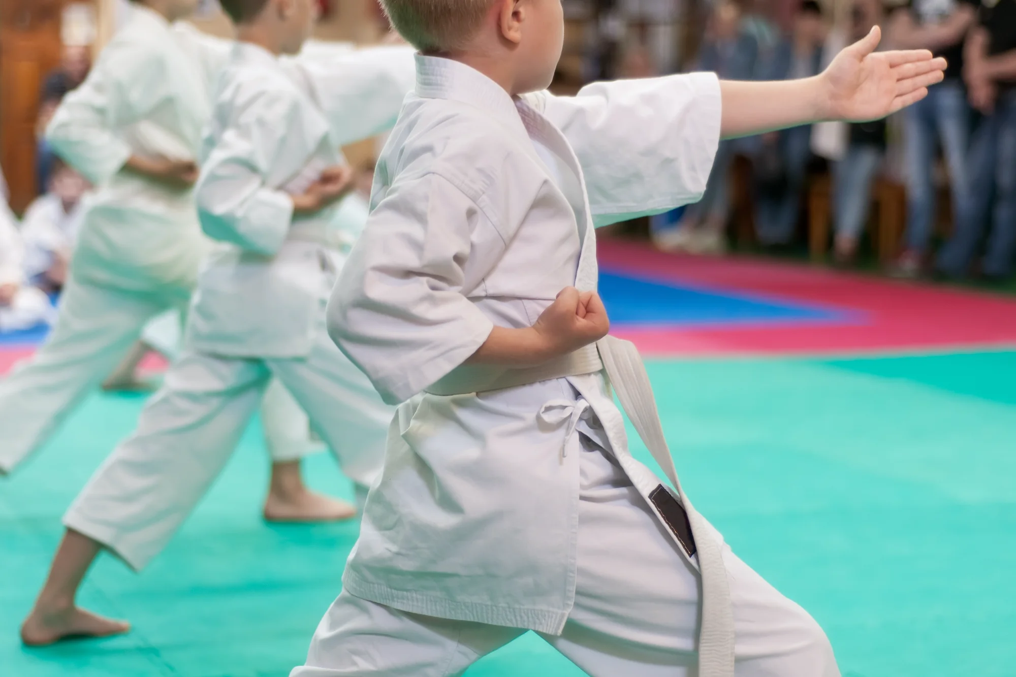 A young martial arts student in a white gi executes a precise stance, holding a focused block with one hand and a fist at the hip.