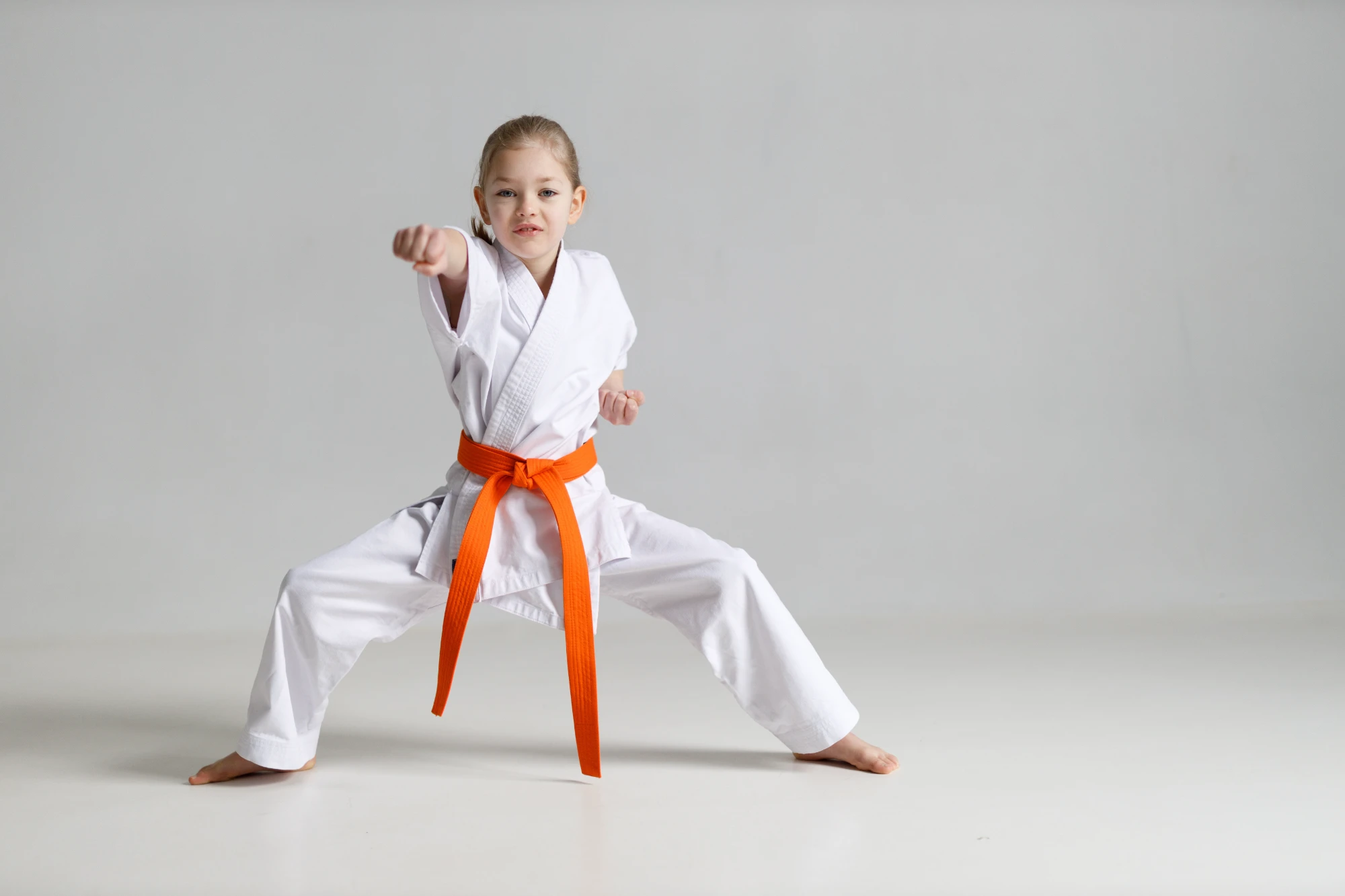 A young girl in a white uniform and orange belt stands in a wide martial arts stance, throwing a punch with a serious expression.
