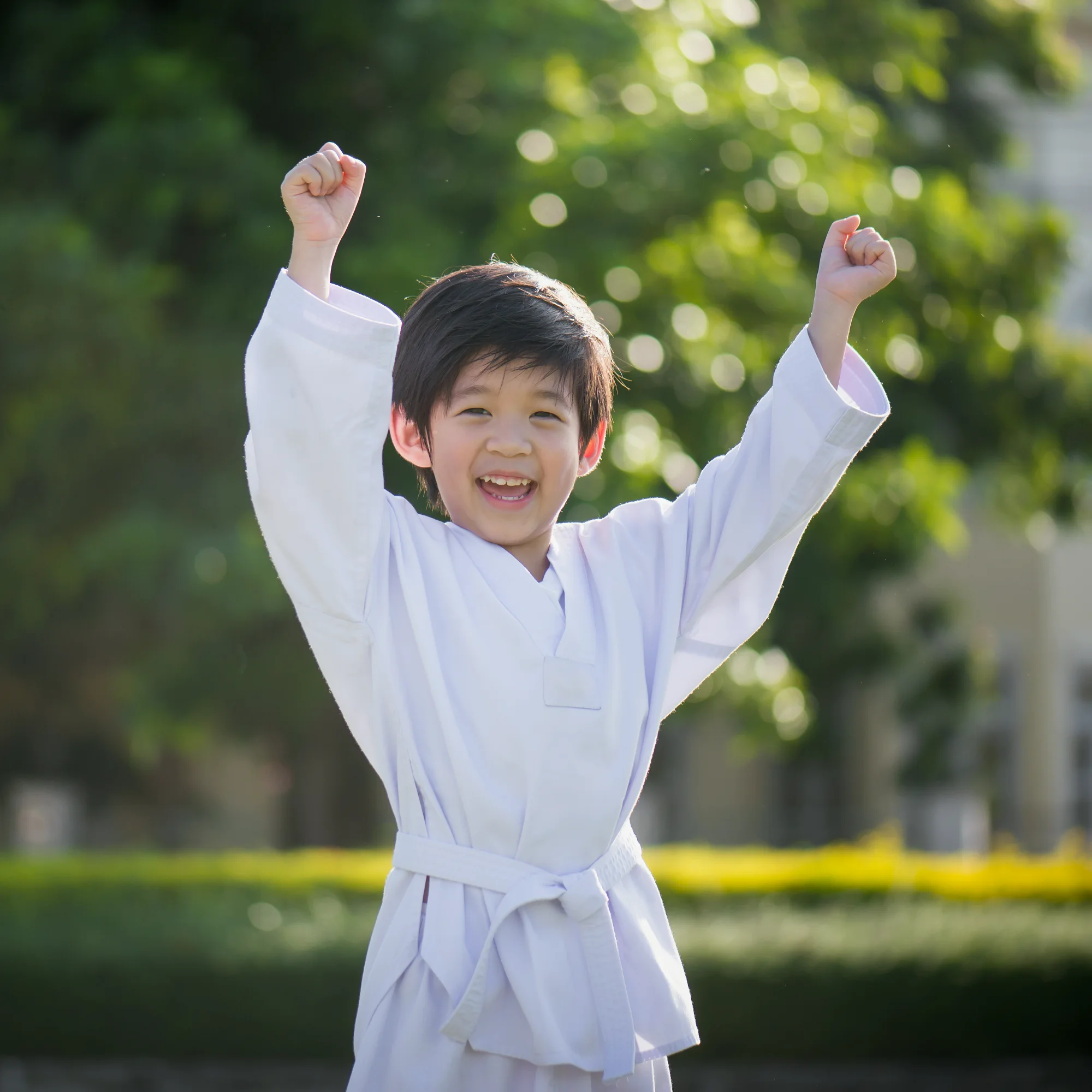 A young student in a white martial arts gi smiles brightly and cheers with both fists raised high outdoors, celebrating success.