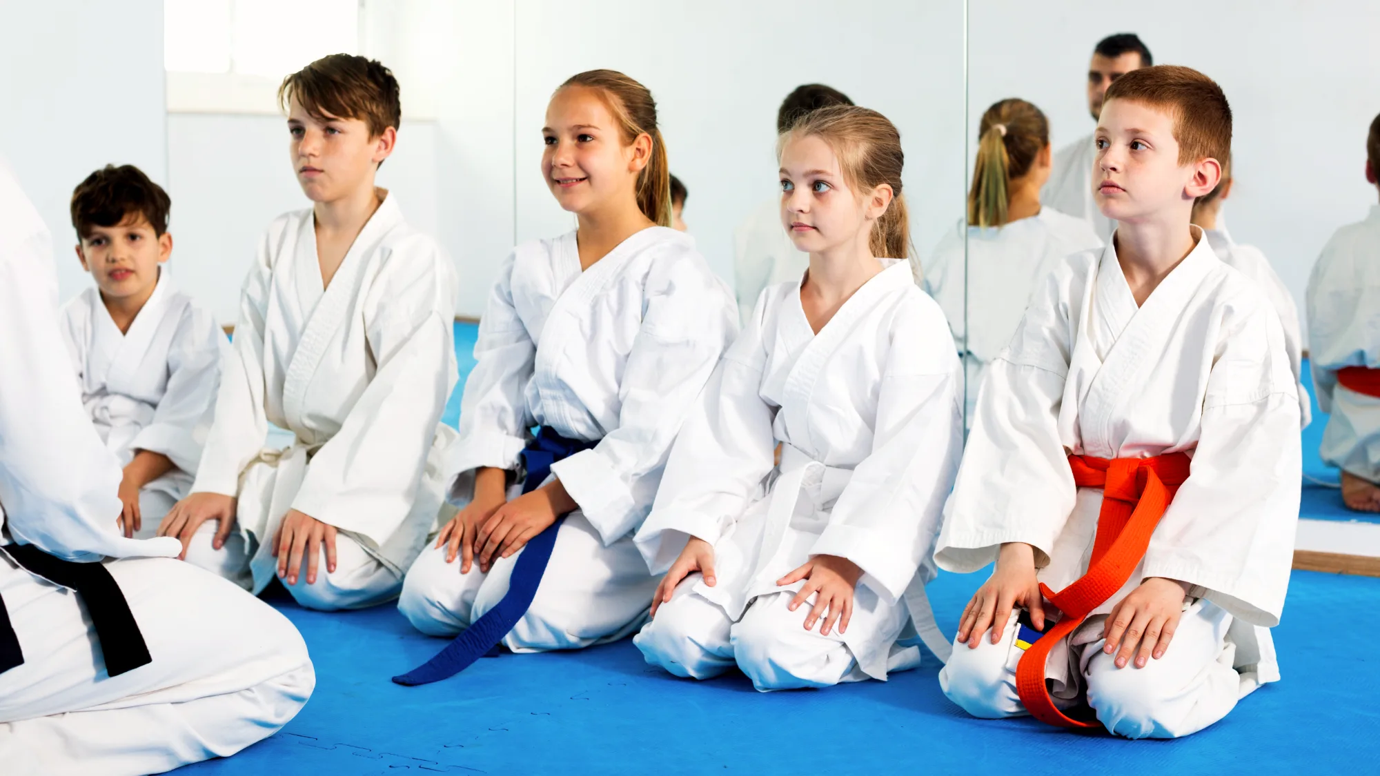 Young karate students in white gis kneel respectfully on a blue mat, looking focused while receiving instruction from their teacher.